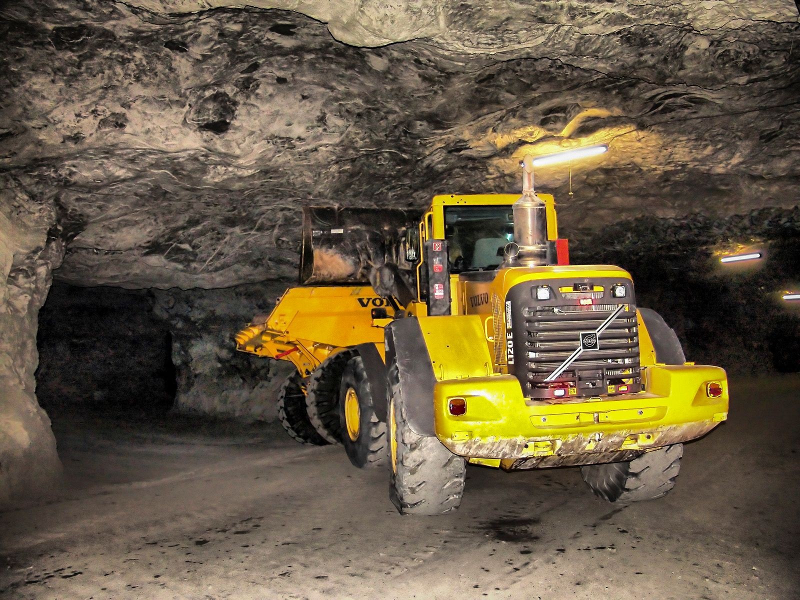 Yellow construction vehicle inside an underground mine tunnel with rugged rock walls and illuminated by ceiling lights.